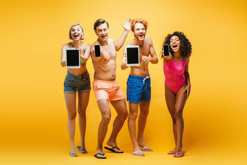 full length view of young multiethnic friends in summer outfit showing gadgets with blank screen on yellow - Photo, Image