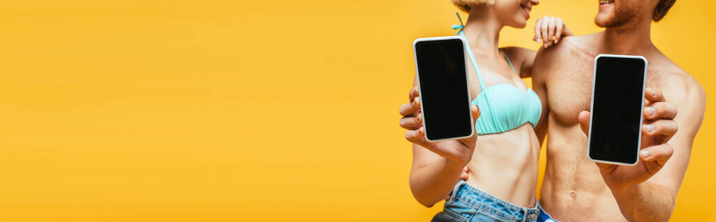 oříznutý pohled na muže a ženu bez trička v plavecké podprsence zobrazující smartphony s prázdnou obrazovkou izolované na žluté, horizontální obraz - Fotografie, Obrázek