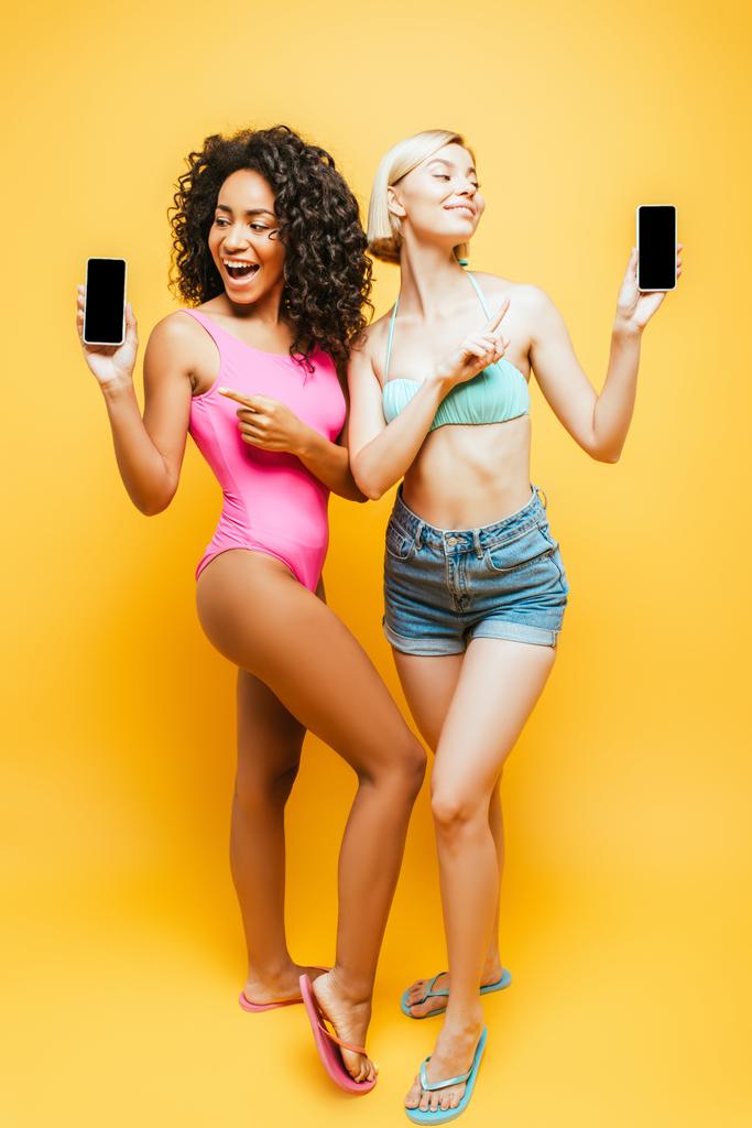 Ganzkörperansicht junger multikultureller Frauen im Sommeroutfit, die auf Smartphones mit leerem Bildschirm auf gelb zeigen - Foto, Bild