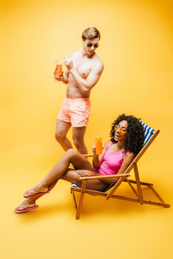jeune femme afro-américaine assise sur une chaise longue avec du jus d'orange près de l'homme torse nu avec verre à cocktail sur jaune - Photo, image