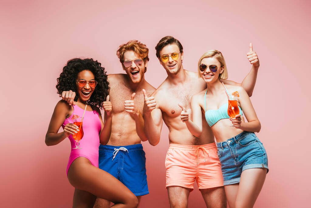 vzrušené multikulturní přátelé ukazující palce nahoru, zatímco drží koktejlové brýle a dívá se na fotoaparát izolované na růžové - Fotografie, Obrázek