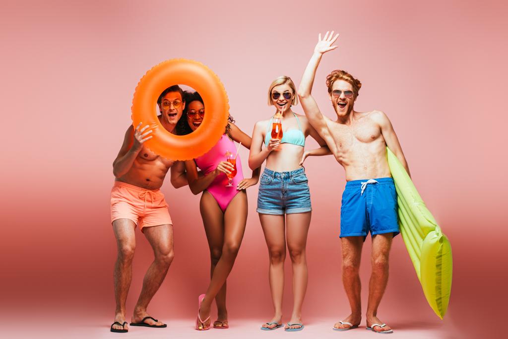 shirtloze man zwaaiende hand in de buurt van opgewonden multiculturele vrienden met zwemring en cocktailglazen geïsoleerd op roze - Foto, afbeelding