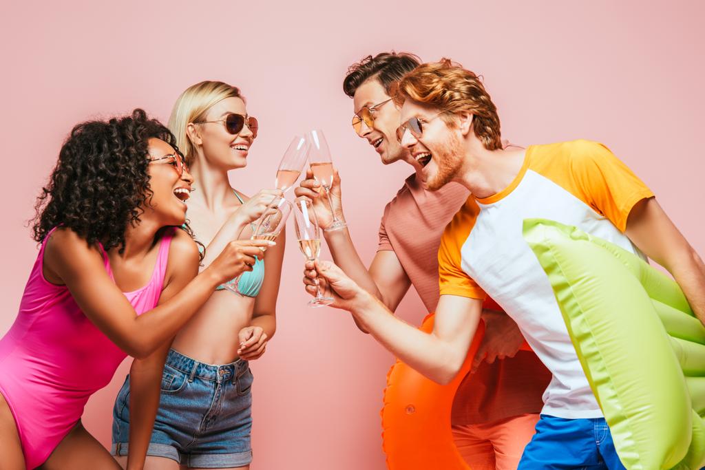 podekscytowani wielokulturowymi przyjaciółmi w letnim stroju i okularach przeciwsłonecznych brzęczących kieliszki szampana na różowym - Zdjęcie, obraz