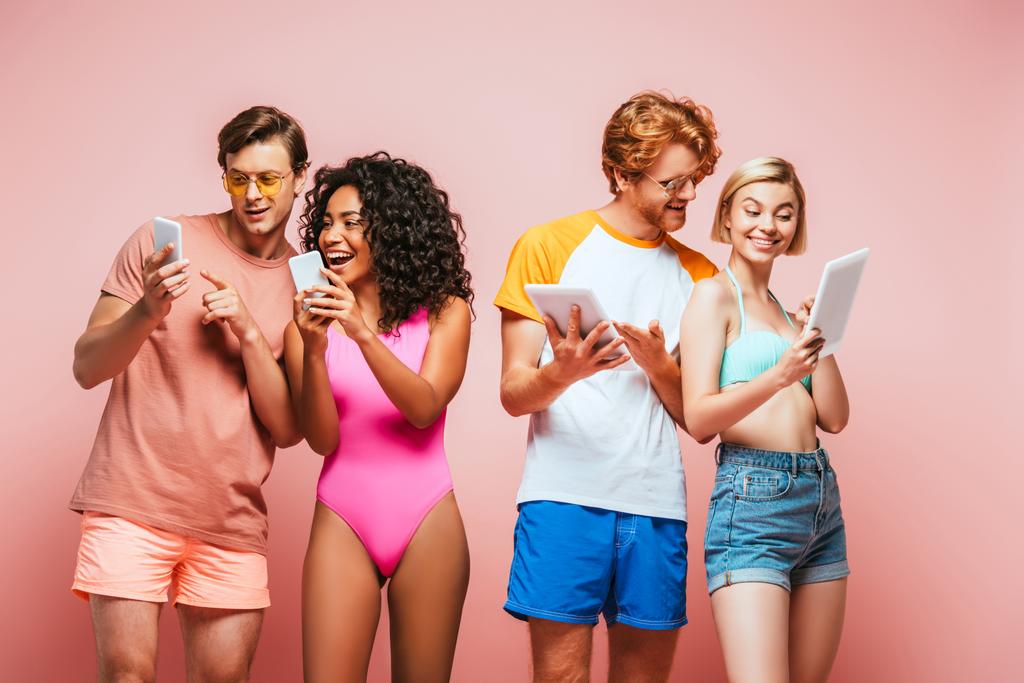 młodzi wielokulturowi przyjaciele w letnim stroju za pomocą cyfrowych urządzeń na różowym - Zdjęcie, obraz