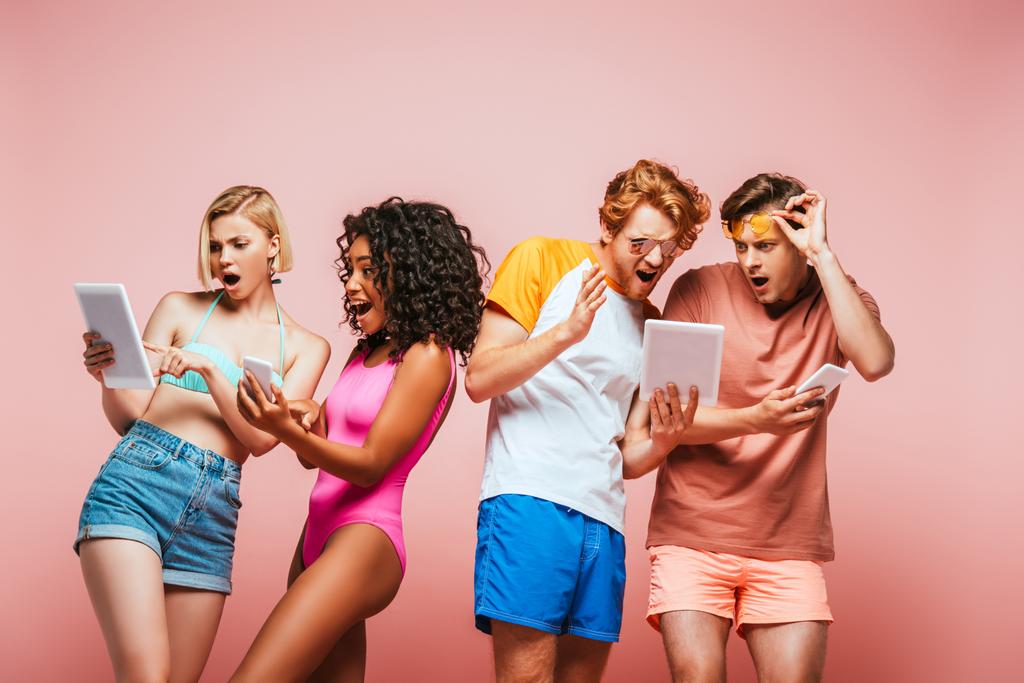 choqué amis multiculturels en tenue d'été en utilisant des appareils numériques isolés sur rose - Photo, image