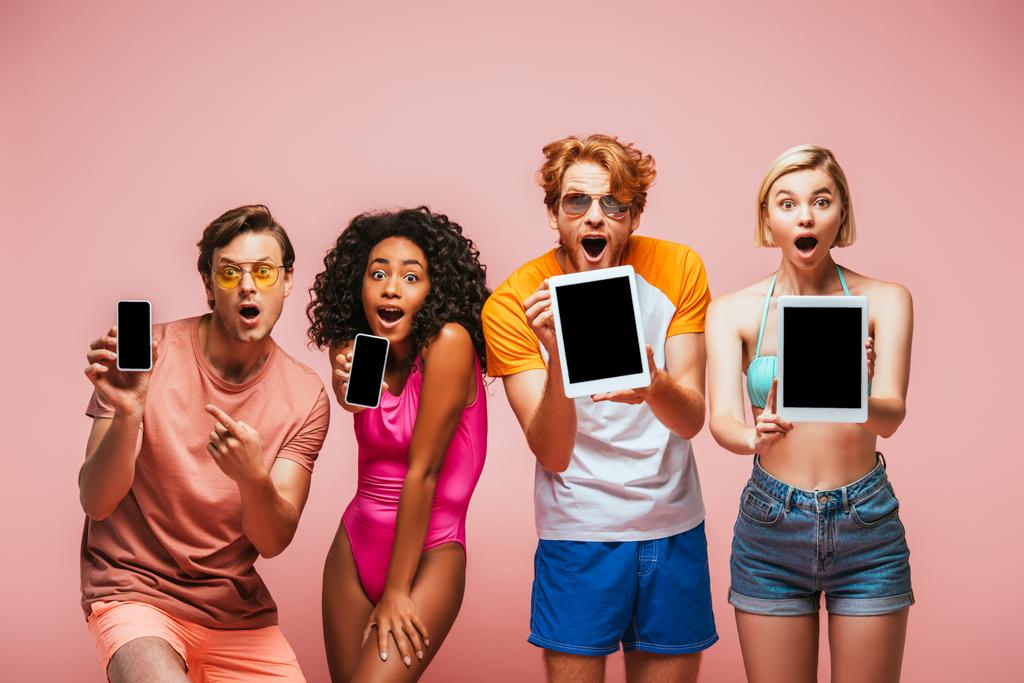ενθουσιασμένοι πολυπολιτισμικοί φίλοι με καλοκαιρινά ρούχα που δείχνουν gadgets με λευκή οθόνη απομονωμένη σε ροζ - Φωτογραφία, εικόνα