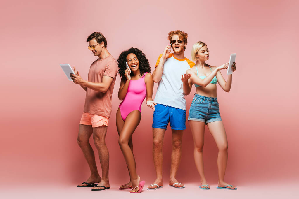 vista completa de los jóvenes amigos multiculturales en traje de verano utilizando dispositivos digitales en rosa - Foto, imagen