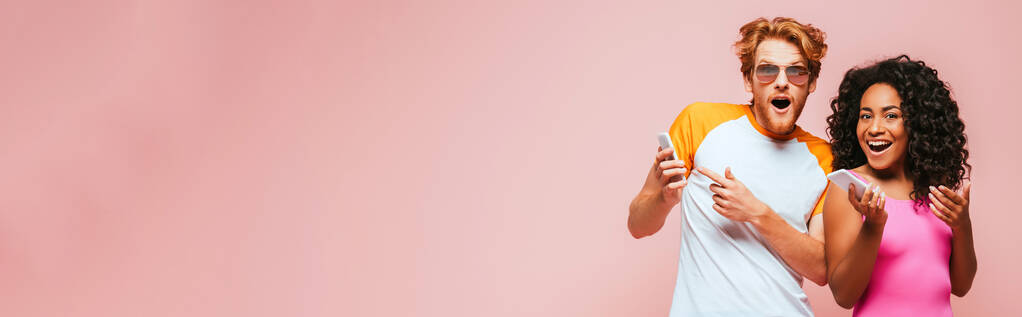 Панорамный снимок многонациональной пары в солнцезащитных очках и купальнике, держащей смартфоны на розовом фоне - Фото, изображение