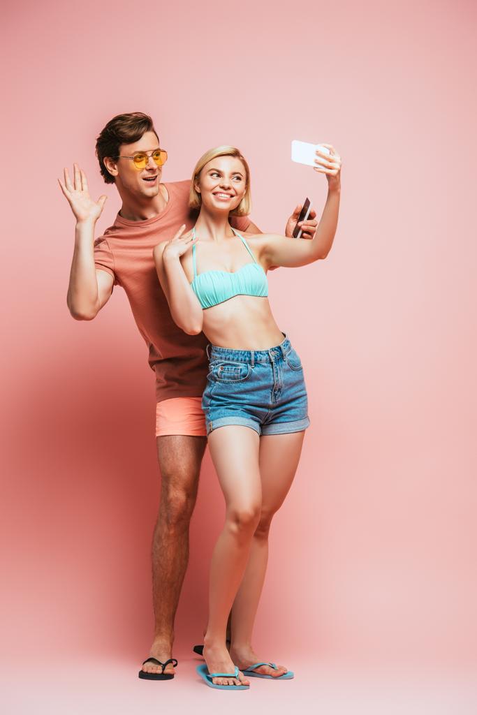 Hombre en gafas de sol que tiene videollamada cerca de la mujer en traje de baño con teléfono inteligente sobre fondo rosa - Foto, imagen