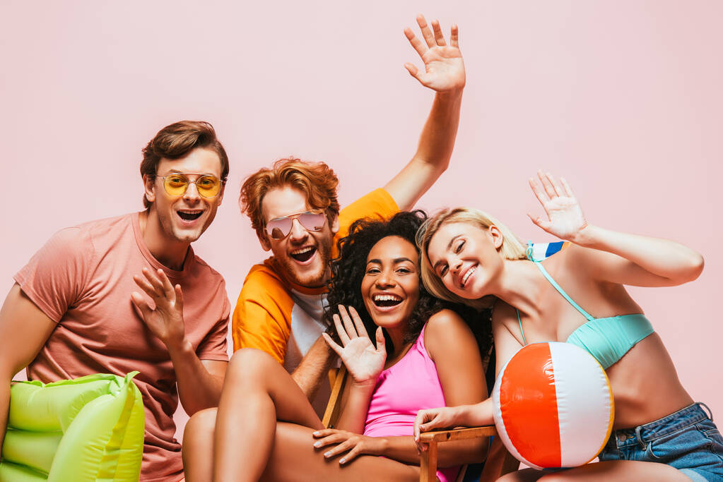 Багатоетнічні друзі в сонцезахисних окулярах з надувним м'ячем і матрацом махають руками на камеру ізольовані на рожевому
  - Фото, зображення