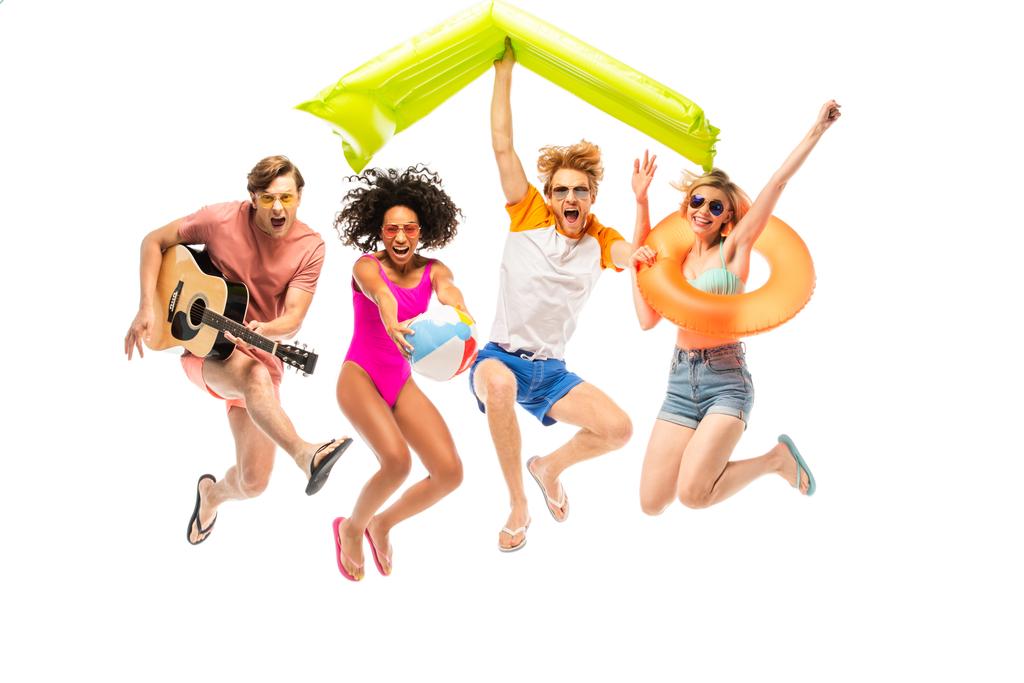 Emocionados amigos multiétnicos con bola inflable, colchón y guitarra acústica saltando aislados en blanco - Foto, imagen