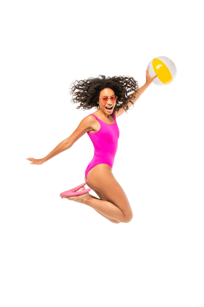 Αφροαμερικανή γυναίκα με γυαλιά ηλίου και μαγιό που κρατάει φουσκωτή μπάλα και πηδάει απομονωμένη στα λευκά - Φωτογραφία, εικόνα
