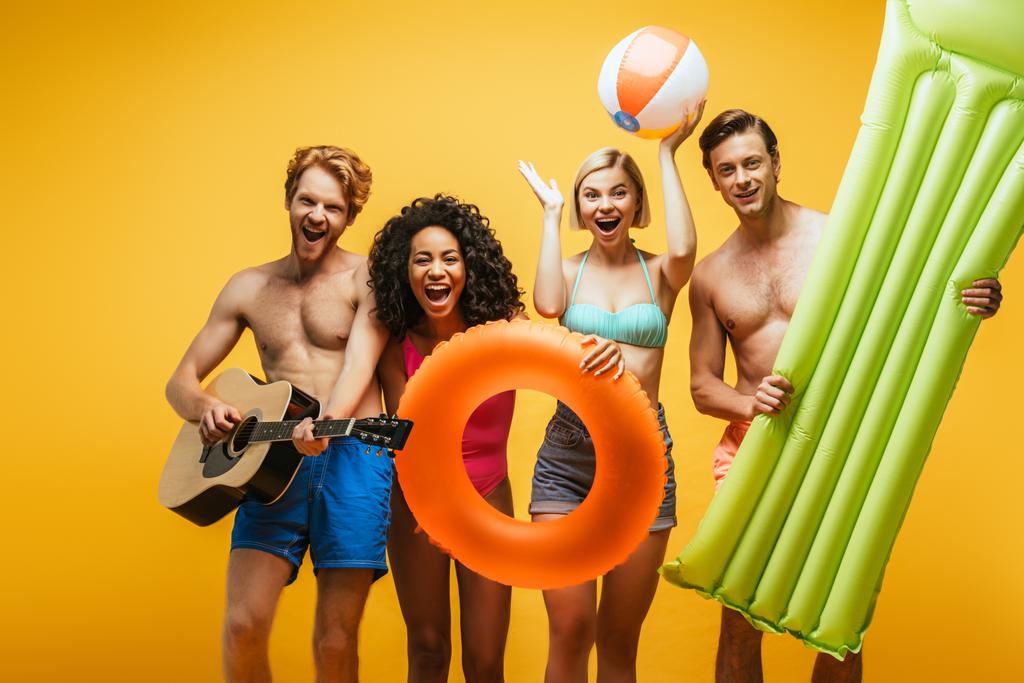 Amigos multiétnicos con guitarra acústica, colchón inflable y bola sobre fondo amarillo - Foto, Imagen