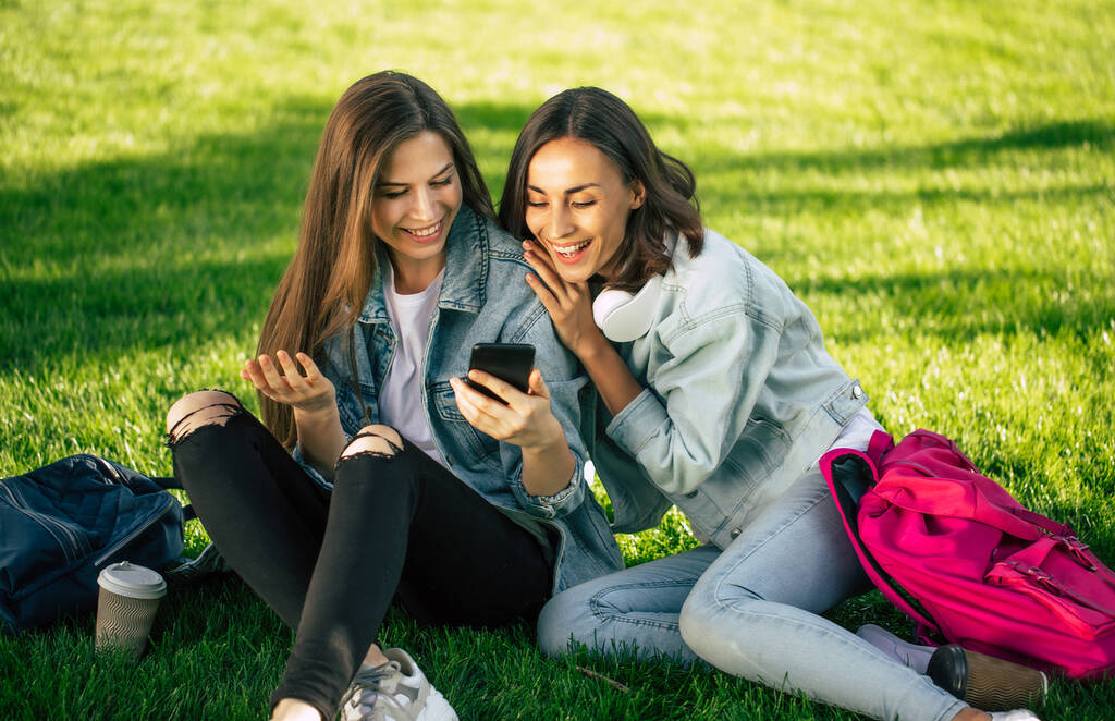 Due amiche studentesse felici ed eccitate in abiti casual in denim si rilassano nel parco universitario con gli smartphone e si divertono. - Foto, immagini