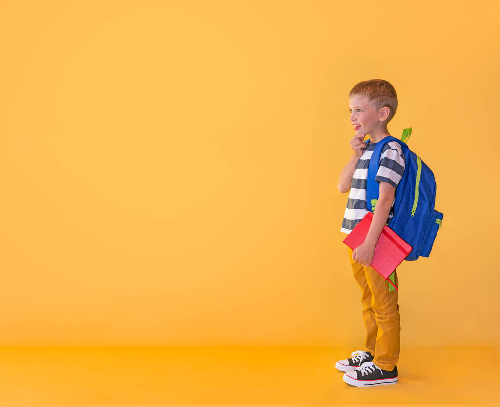 Zainteresowany chłopiec z książką i plecakiem patrzy w odstęp na żółtym tle. Koncepcja powrotu do szkoły. - Zdjęcie, obraz