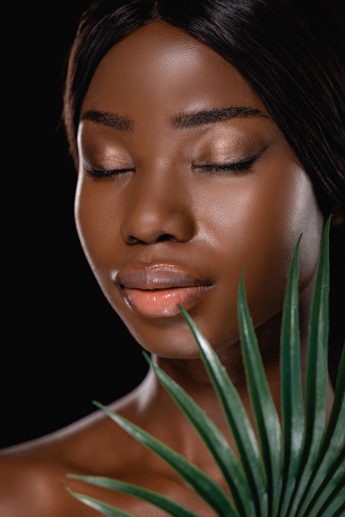mujer desnuda afroamericana con los ojos cerrados y hoja de palma verde aislado en negro - Foto, imagen