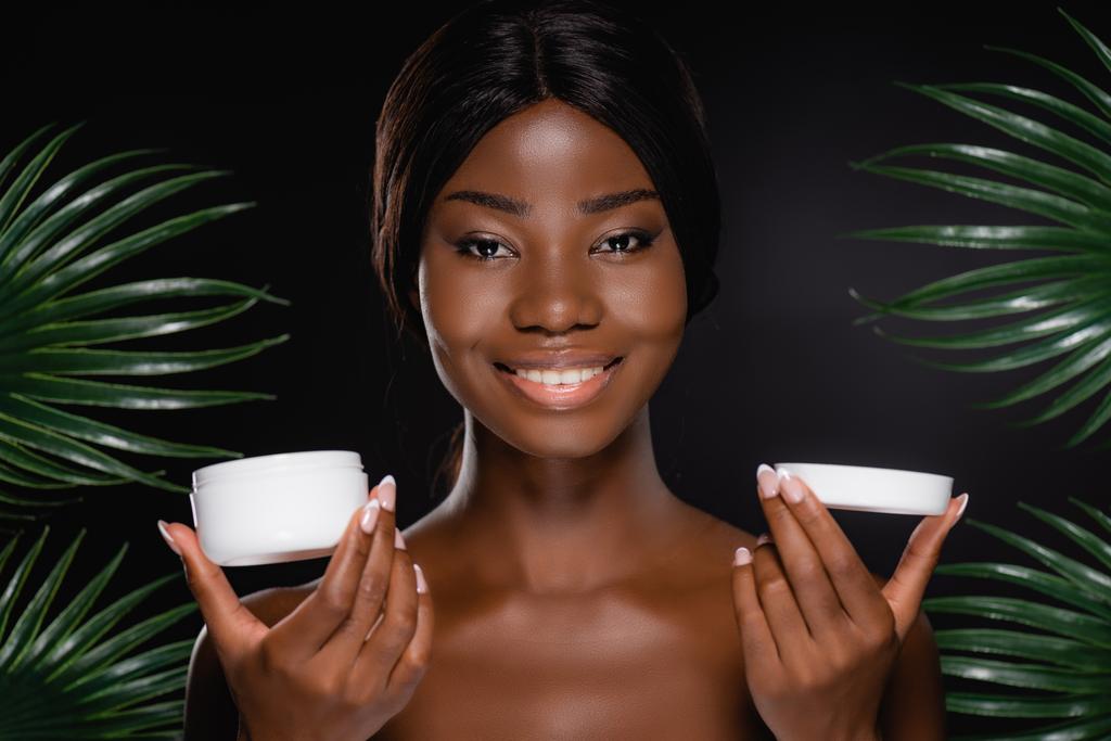 Siyah palmiye yapraklarının yanında kozmetik kremi olan Afro-Amerikan çıplak kadın. - Fotoğraf, Görsel