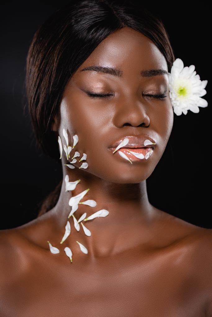 nackte afrikanisch-amerikanische Frau mit geschlossenen Augen, weiße Chrysantheme im Haar und Blütenblätter am Hals isoliert auf schwarz - Foto, Bild