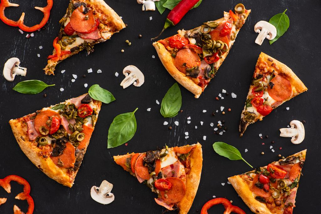 вид сверху на вкусные итальянские ломтики пиццы с овощами и салями на черном фоне - Фото, изображение