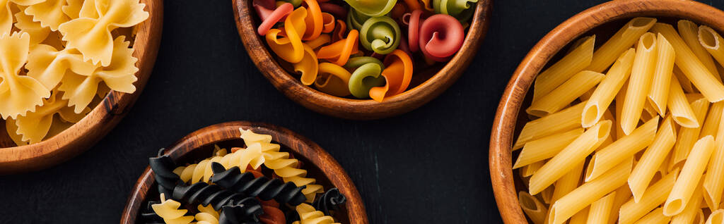 top view of assorted colorful Italian pasta in wooden bowls on black background, panoramic shot - Photo, Image