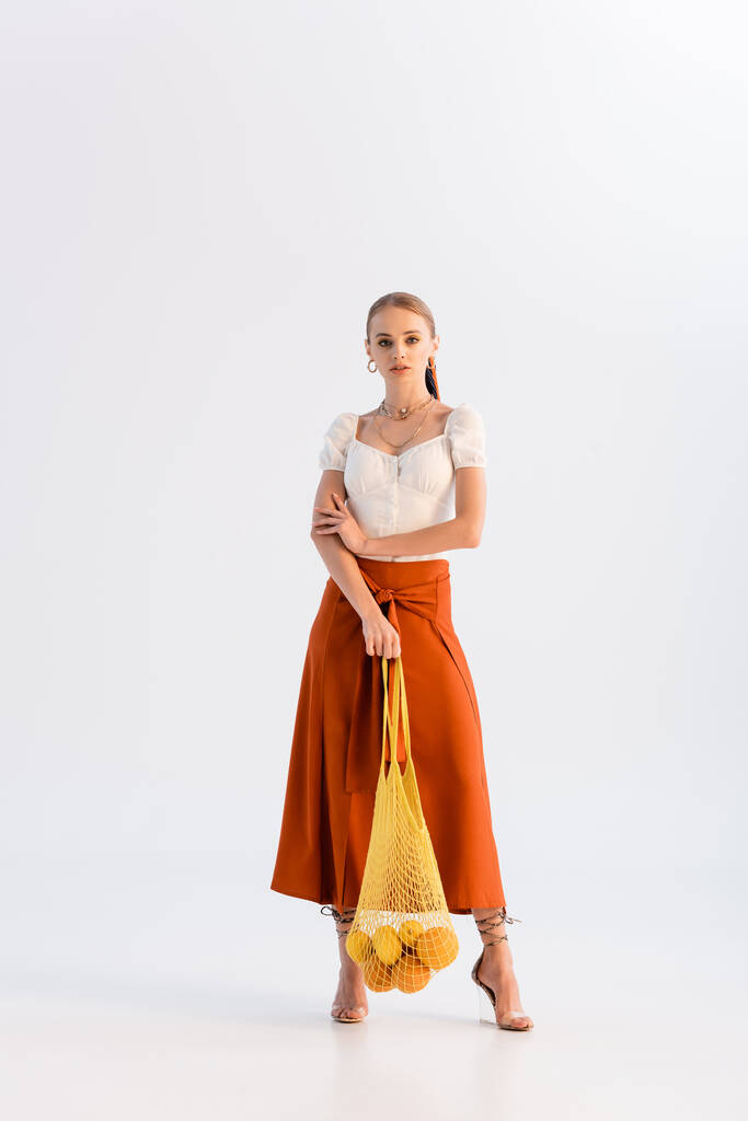 visão de comprimento total da mulher na moda posando com frutas cítricas em saco de cordas isolado no branco - Foto, Imagem