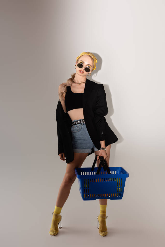 teljes hosszúságú kilátás divatos nő pózol kék bevásárló kosár szürke háttér - Fotó, kép