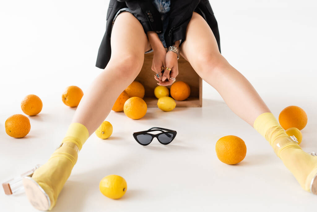 vista recortada de la mujer de moda en calcetines amarillos y sandalias posando en caja de madera cerca de cítricos dispersos sobre fondo blanco - Foto, Imagen