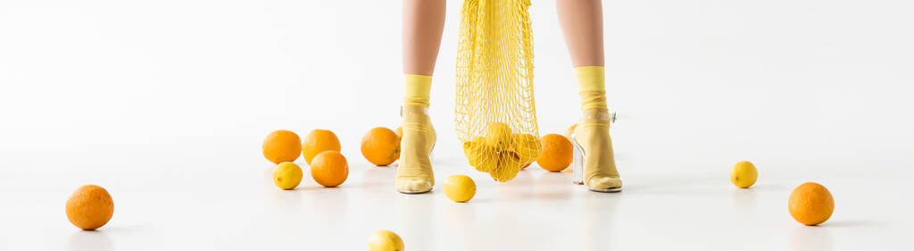 vista recortada de las piernas femeninas en calcetines y sandalias amarillas y bolsa de cuerdas cerca de cítricos dispersos sobre fondo blanco, plano panorámico - Foto, Imagen