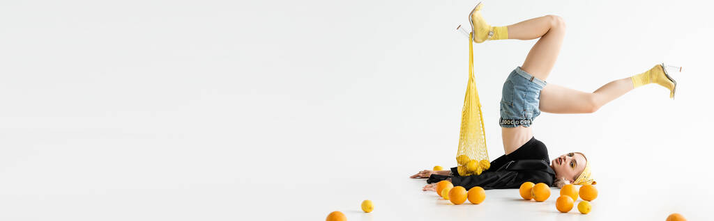 visão lateral da mulher na moda posando com saco de cordas na perna perto de frutas cítricas espalhadas no fundo branco, tiro panorâmico - Foto, Imagem