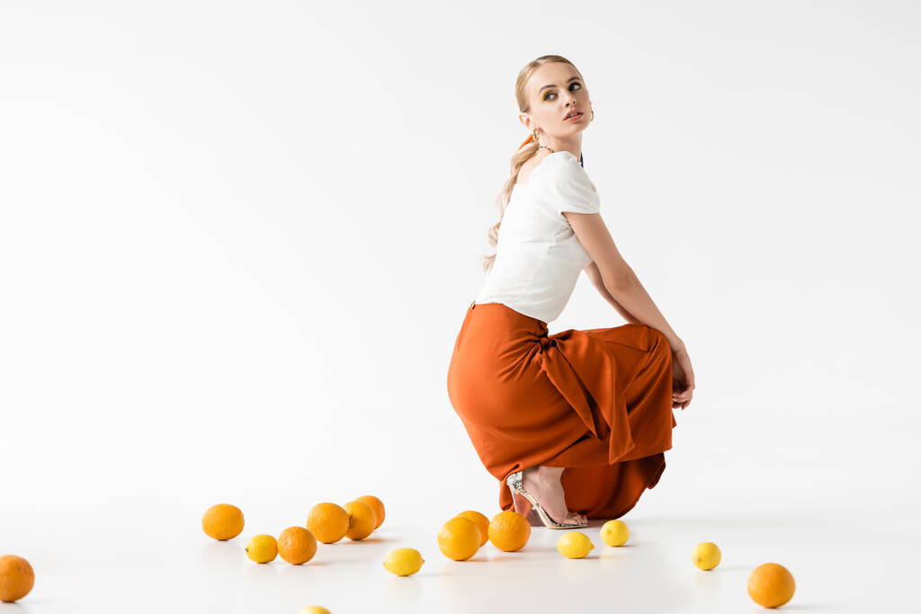 vista lateral da mulher loira elegante posando perto de frutas cítricas no fundo branco - Foto, Imagem