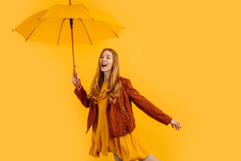 Ευτυχισμένο κορίτσι με κίτρινο φόρεμα και φθινοπωρινό μπουφάν, γελώντας και διασκεδάζοντας με κίτρινη ομπρέλα, στέκεται σε κίτρινο φόντο - Φωτογραφία, εικόνα