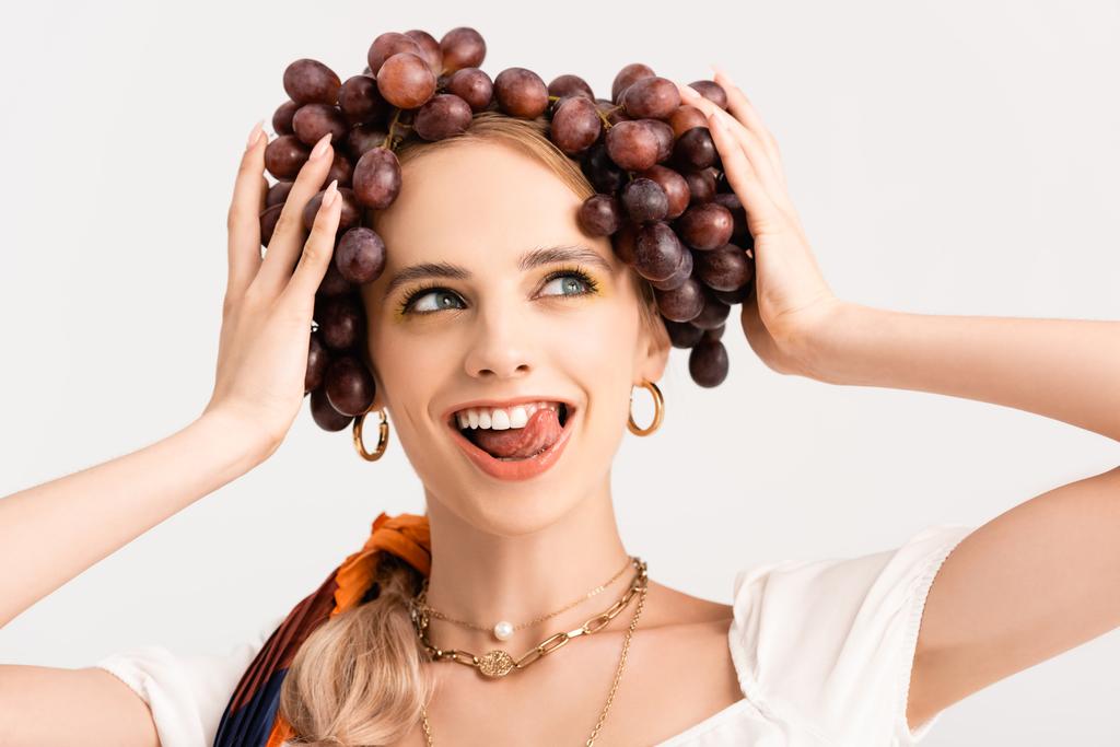деревенская блондинка держит виноград на голове и показывает язык изолирован на белом - Фото, изображение