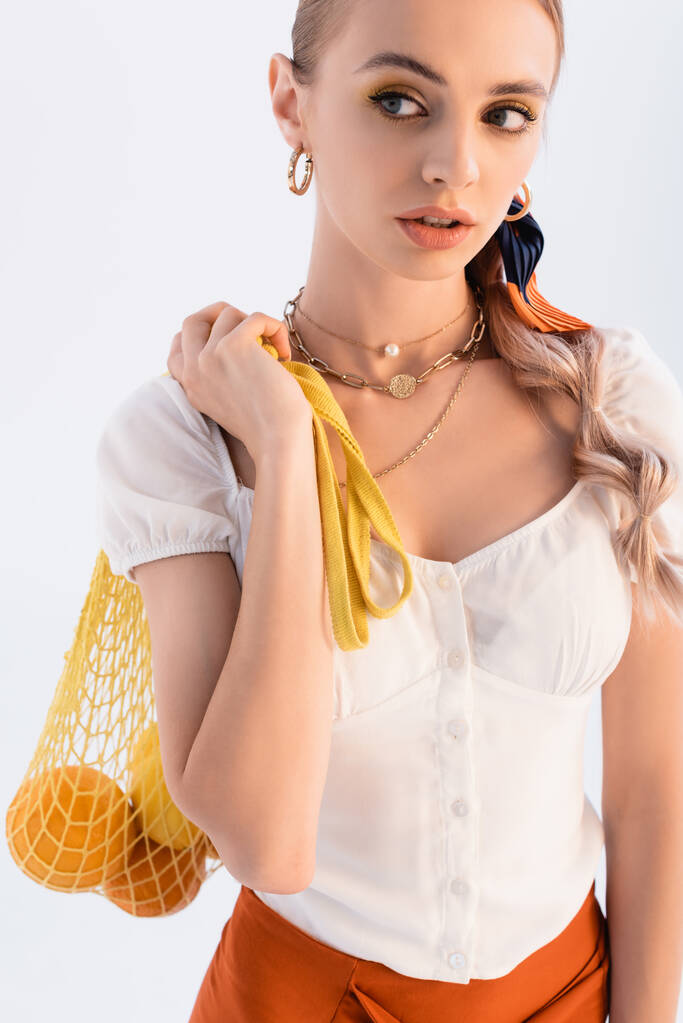 mulher loira rústica posando com frutas cítricas em saco de corda amarela e olhando para longe isolado no branco - Foto, Imagem