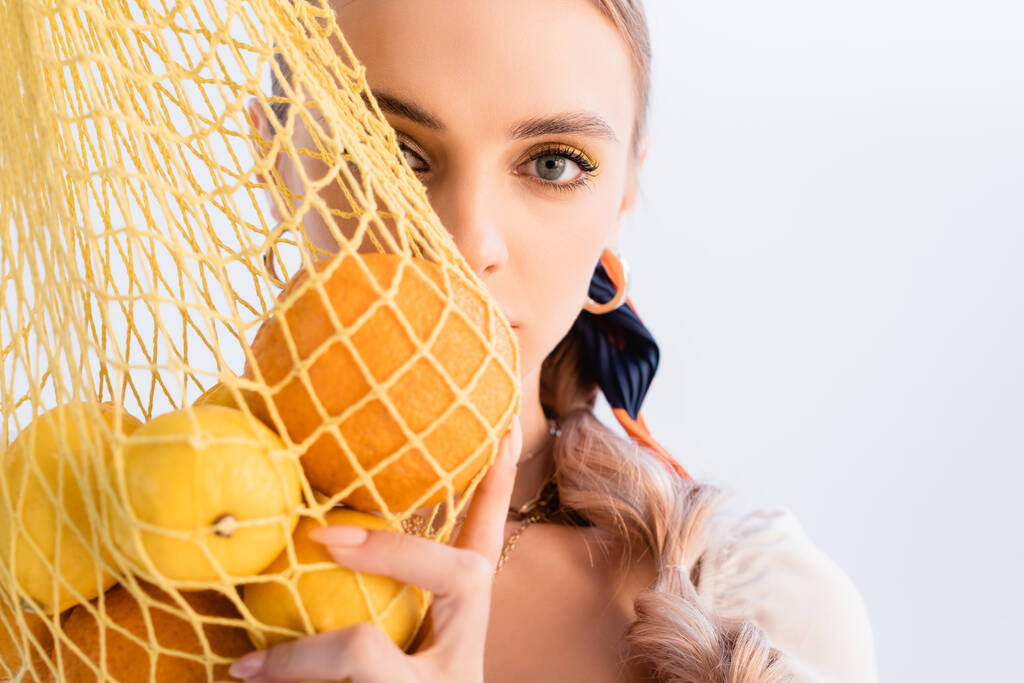 Rustic blonde woman posing with citrus fruits in yellow string bag in front of face isolated on white - Фото, изображение