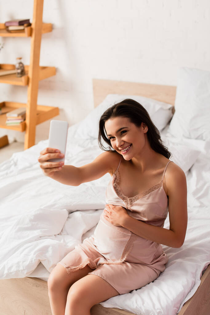 mujer joven embarazada sentada en la cama y tomando selfie en el dormitorio - Foto, Imagen