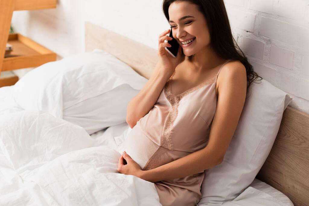 νεαρή και έγκυος γυναίκα που μιλάει στο smartphone στο υπνοδωμάτιο   - Φωτογραφία, εικόνα