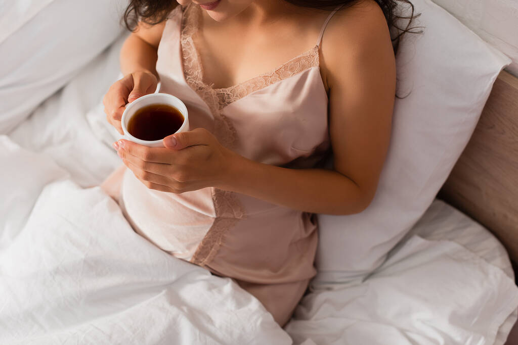 abgeschnittene Ansicht einer Frau im seidenen Nachthemd, die eine Tasse Tee hält  - Foto, Bild