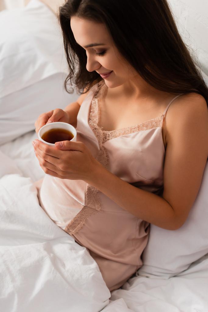 έγκυος νεαρή γυναίκα σε μεταξωτό νυχτικό κρατώντας φλιτζάνι τσάι  - Φωτογραφία, εικόνα