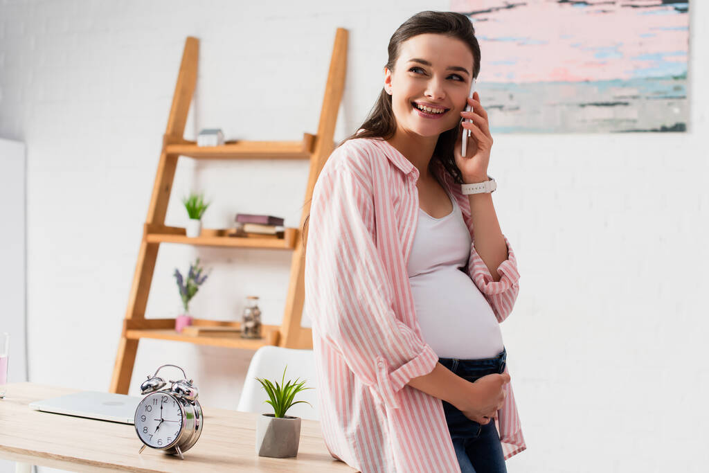 mulher grávida falando no smartphone e de pé perto da mesa com despertador vintage e planta - Foto, Imagem