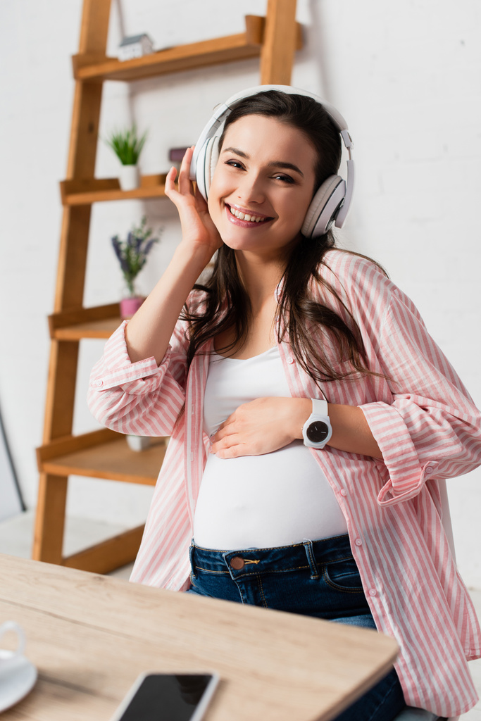 szelektív fókusz a terhes nő vezeték nélküli fejhallgató zenét hallgatni közelében okostelefon üres képernyőn  - Fotó, kép