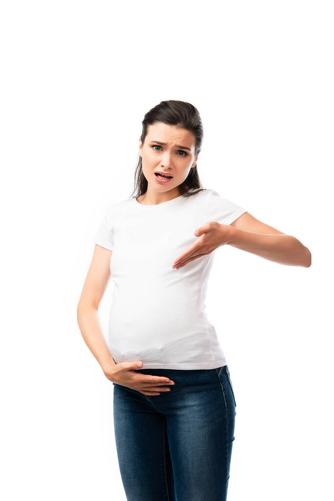 mujer embarazada joven en camiseta blanca apuntando con la mano hacia el vientre aislado en blanco  - Foto, Imagen