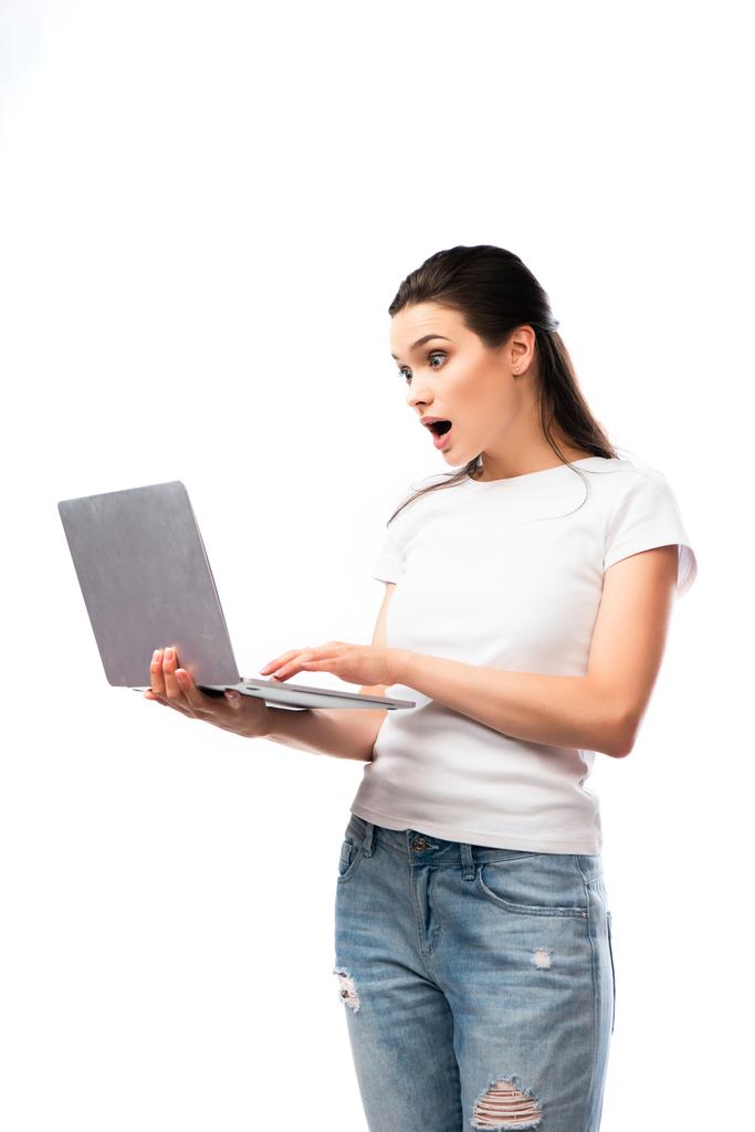 σοκαρισμένη μελαχρινή γυναίκα σε λευκό t-shirt χρησιμοποιώντας φορητό υπολογιστή απομονωμένο σε λευκό  - Φωτογραφία, εικόνα