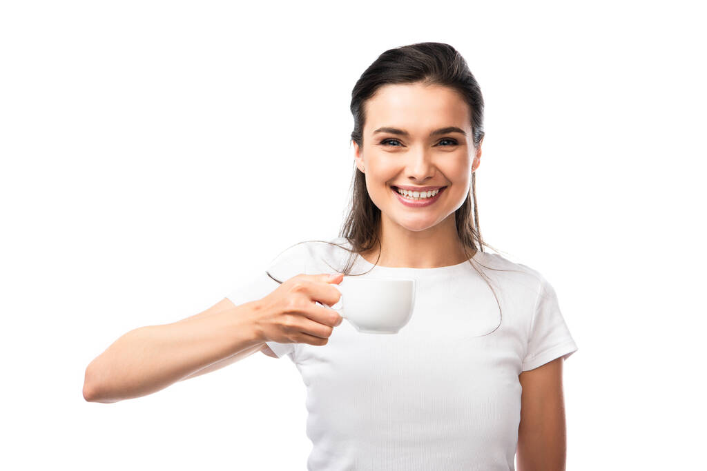 femme brune en t-shirt blanc regardant la caméra et tenant tasse avec café isolé sur blanc  - Photo, image