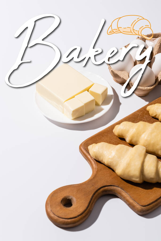 croissants crus sur planche à découper en bois près du beurre, oeufs et lettrage boulangerie sur fond blanc - Photo, image