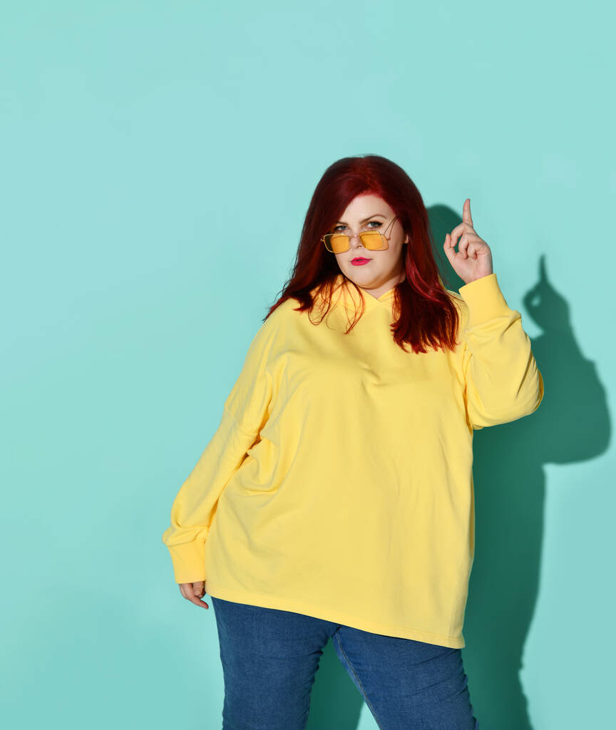 Serious plus-size woman in stylish casual attire and glasses standing in a confident pose holding finger up in eureka gesture, isolated on blue - Photo, Image