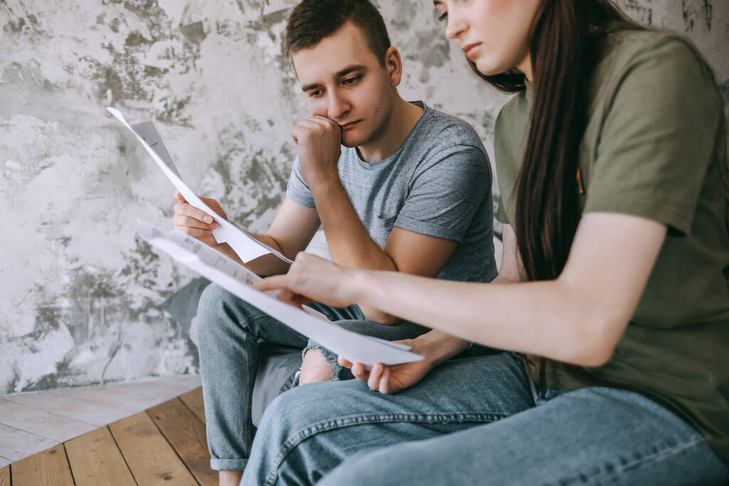 A young couple manages finances by looking through their bank accounts in the living room by the bed. Woman and man look at documents together. Planning budget expenses. High quality photo - Photo, Image