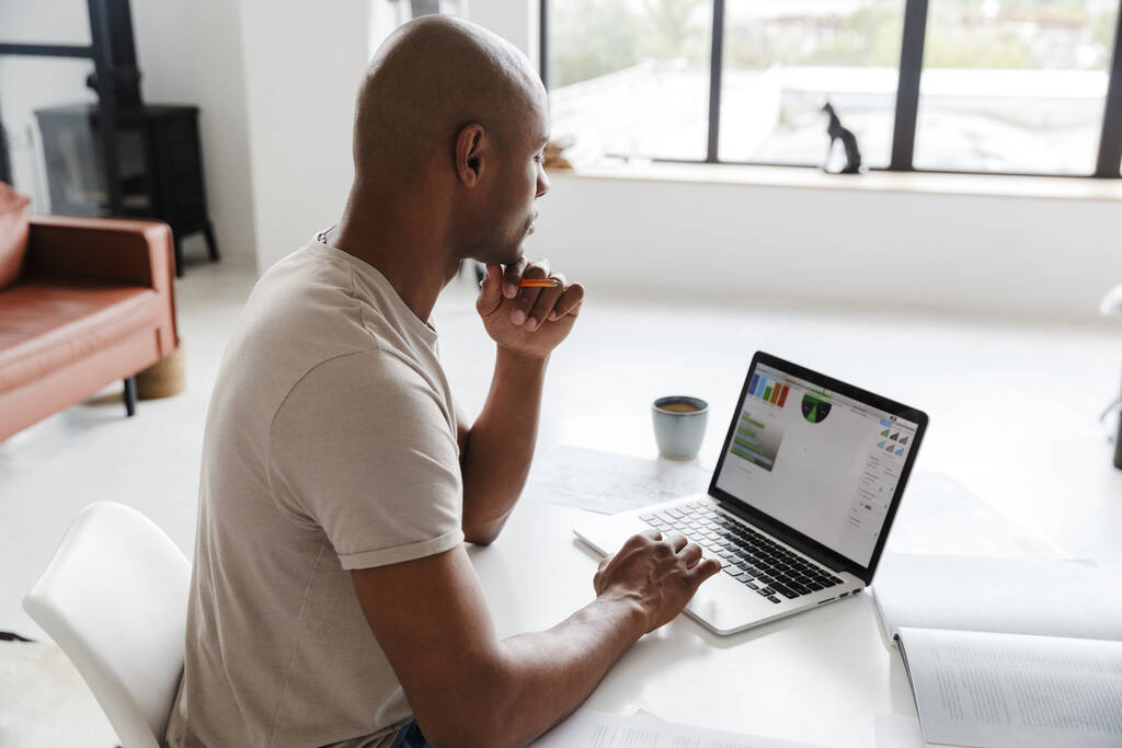Kuva keskittynyt afrikkalainen amerikkalainen mies työskentelee laptop istuessaan pöydässä olohuoneessa - Valokuva, kuva