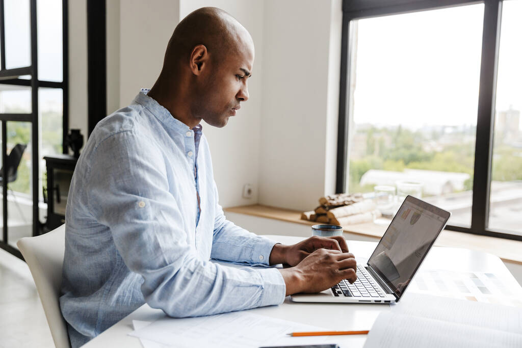 Фото сконцентрированного африканского американца, работающего с ноутбуком, сидящего за столом в гостиной - Фото, изображение