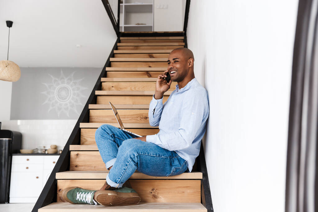 Foto eines lächelnden afrikanisch-amerikanischen Mannes, der Laptop benutzt und auf dem Handy spricht, während er zu Hause auf der Treppe sitzt - Foto, Bild