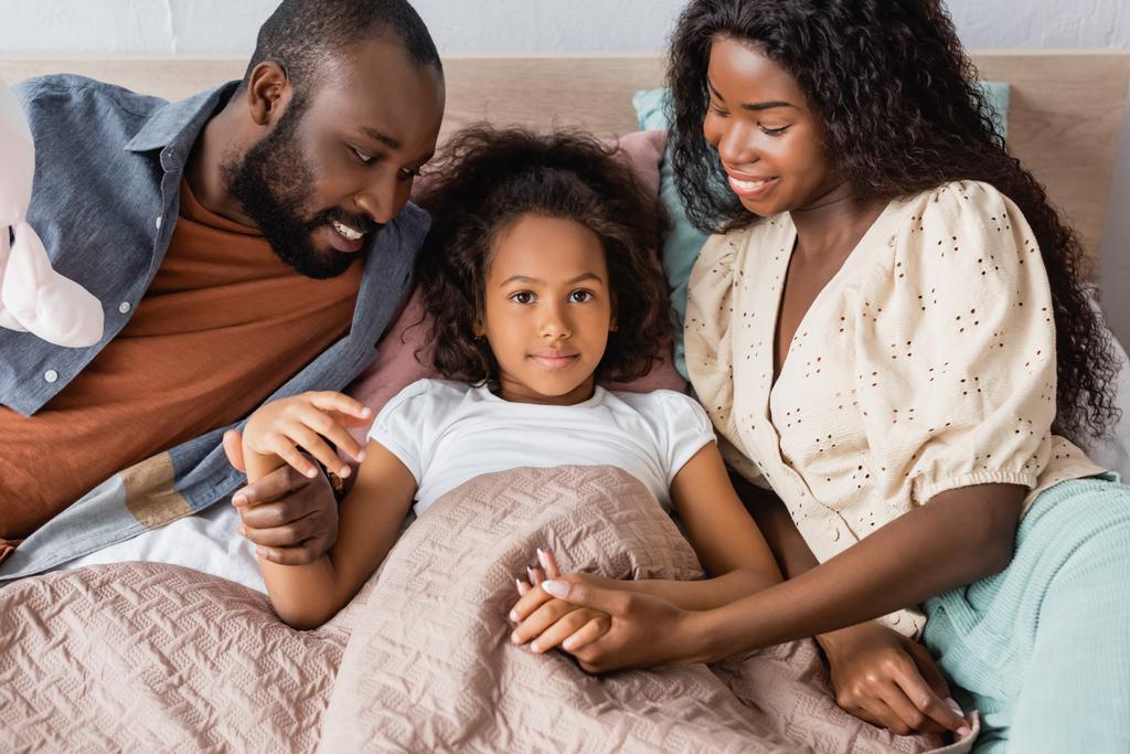 hoge hoek uitzicht van Afrikaans Amerikaans meisje liggend in bed en kijken naar camera terwijl ouders zitten in de buurt en aanraken van haar handen - Foto, afbeelding